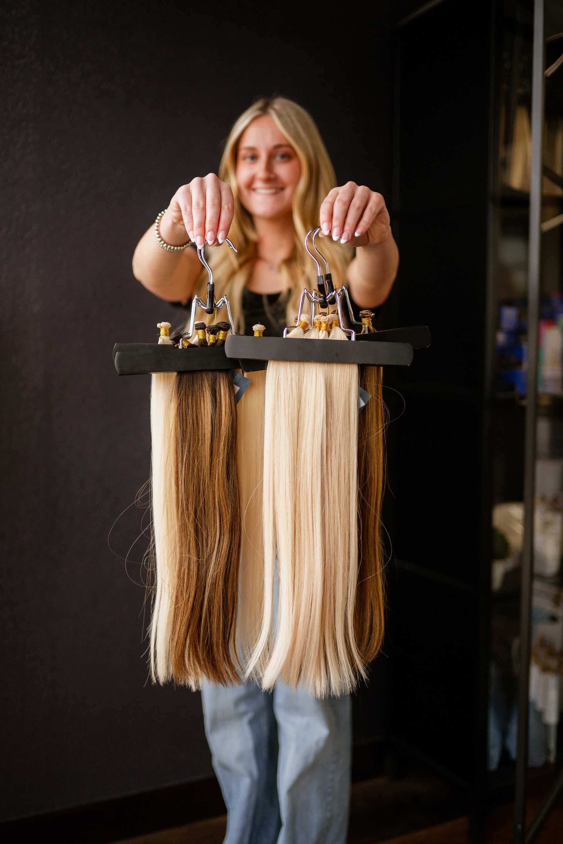 A woman holds two sets of hair extensions, one blonde and one brown, smiling in a salon setting.